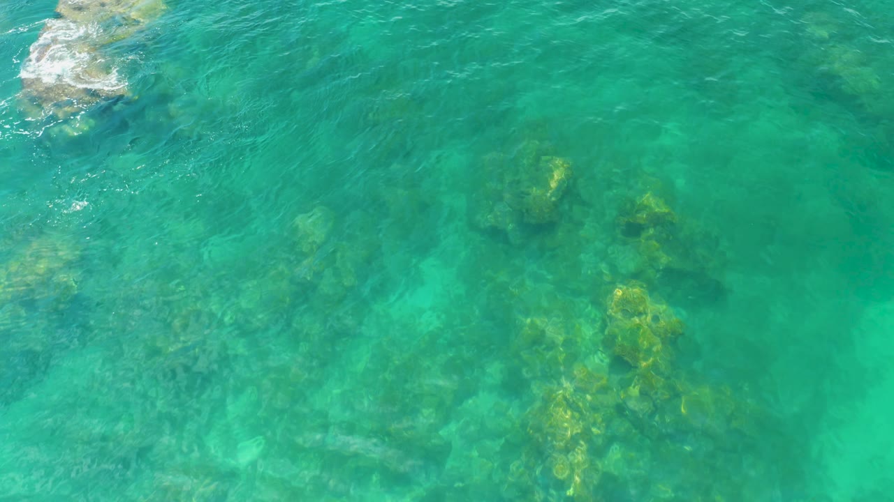 An overhead Palmilla Beach frame shows bright turquoise water, darker reef patches below, and a small rock with white foam near the top, no visible people.