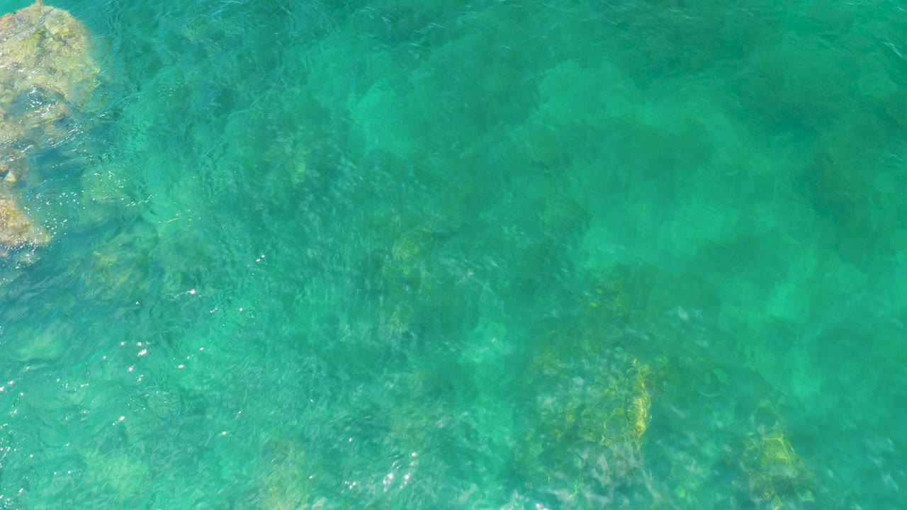 Overhead view of turquoise water at Palmilla Beach, Baja California Sur, with a pale rock formation along the left edge and no visible people.