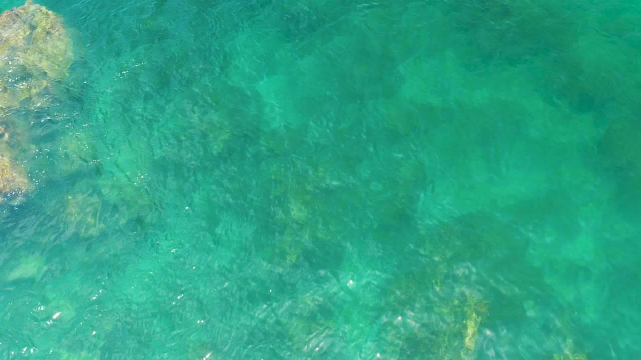 Aerial frame over Palmilla Beach water, with a larger rock formation entering from the upper left and darker submerged patches below, no visible people.
