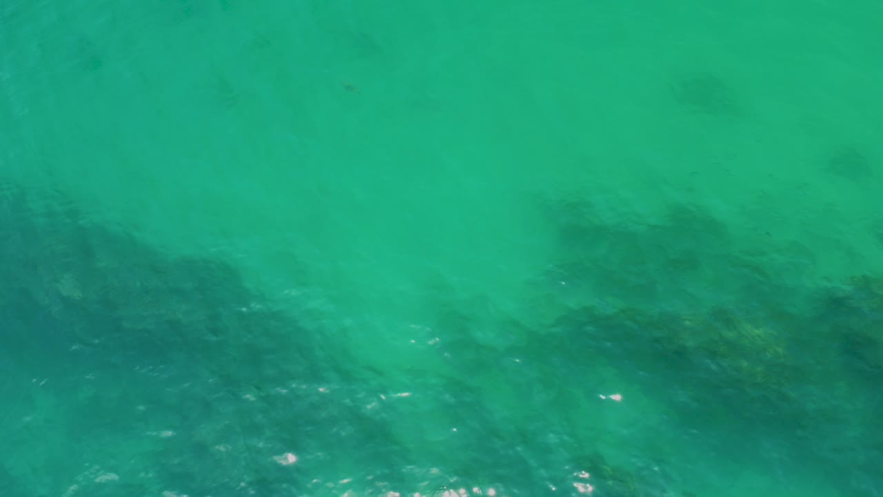 Overhead view of turquoise water at Palmilla Beach with dark submerged rock patches near the lower edge and no visible people.