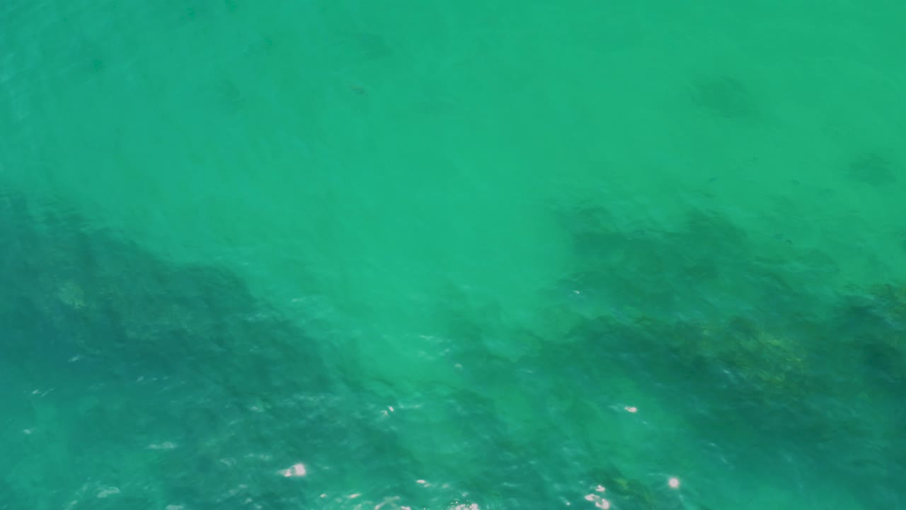 Clear green-turquoise water at Palmilla Beach covers a long dark rock band across the lower half, with small bright reflections and no visible people.