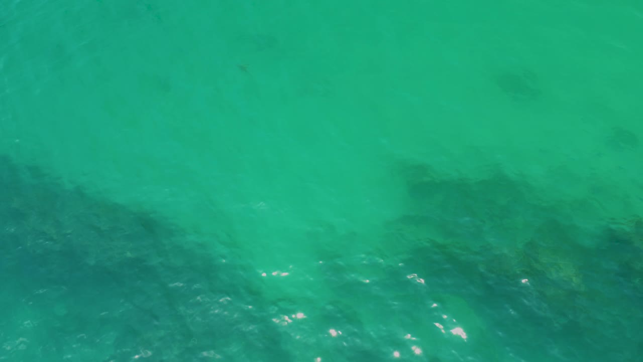 A Palmilla Beach overhead frame shows shallow turquoise water with darker submerged rocks running along the bottom of the frame and no visible people.