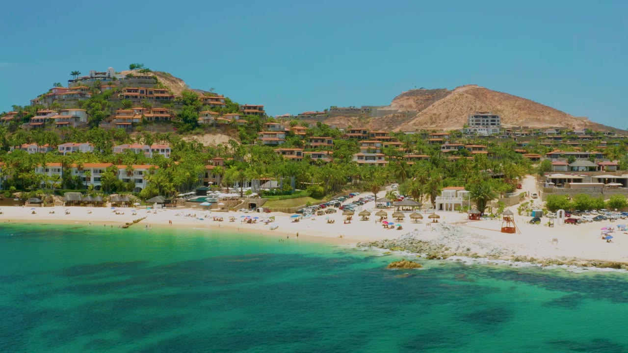 Aerial frame over Palmilla Beach, Baja California Sur, showing turquoise water in front, a pale sand beach with umbrellas, palm trees, hillside homes, and no visible people.