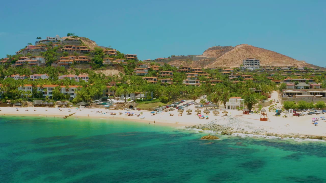 Early aerial view of Palmilla Beach with green-blue water across the foreground, rows of beach umbrellas near the sand, hillside residences behind palms, and no visible people.