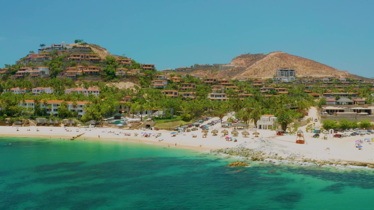 Aerial frame at Palmilla Beach showing turquoise shallows, bright sand, umbrella rows, palms, hillside homes, dry hills, and no visible people.