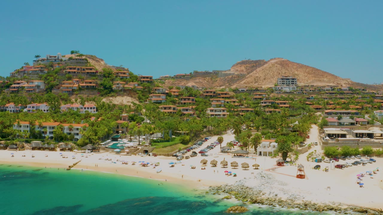 Opening frame over Palmilla Beach in Baja California Sur, with turquoise water at lower left, pale sand, umbrellas, palms, hillside houses, dry mountains, and no visible people.