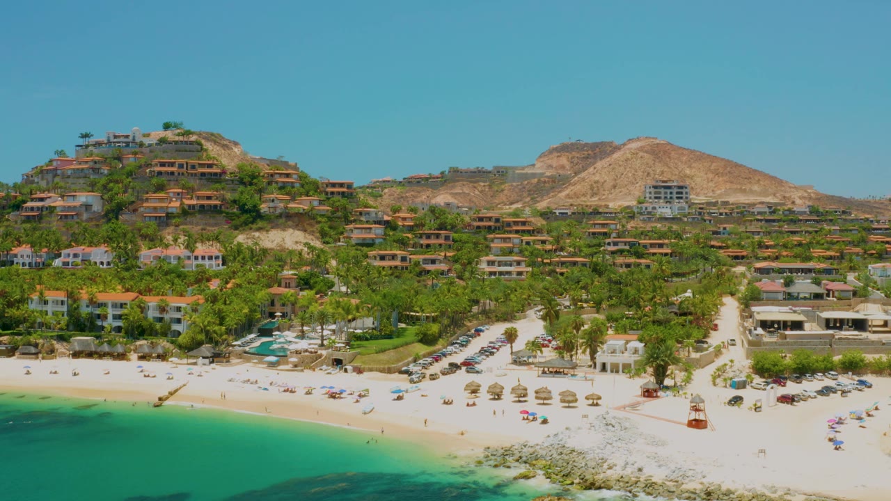 Elevated Palmilla Beach frame with turquoise water across the lower left, a broad sand strip with small umbrella rows, palms and buildings behind it, dry hills, and no visible people.
