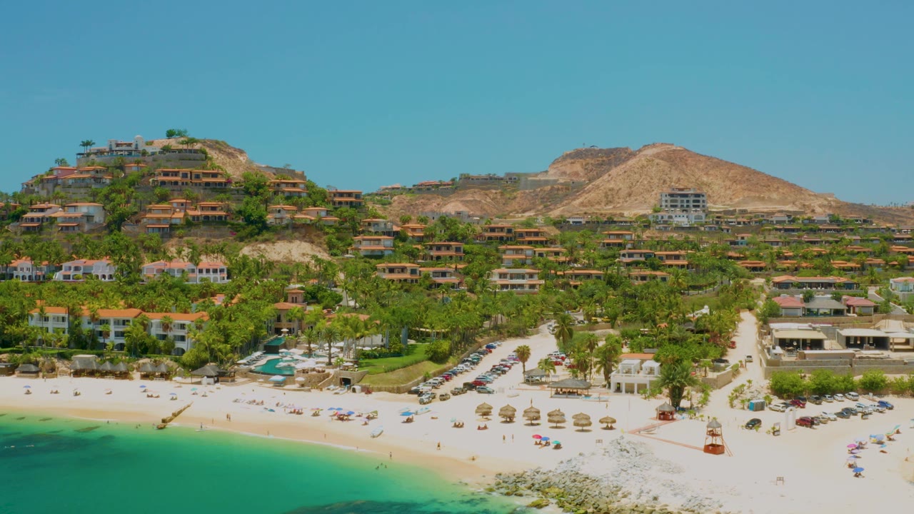 Aerial beach scene with turquoise sea, white sand, palms, parked cars, homes, and hills.