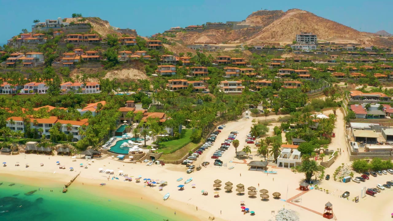 Palmilla Beach aerial still with a curved turquoise shoreline at lower left, straw umbrellas on the sand, a central access road, hillside residences, and no visible people.