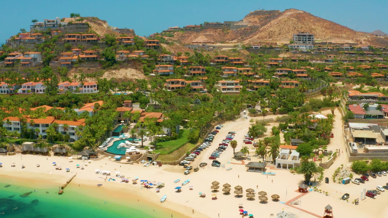 Wide aerial view at Palmilla Beach, Baja California Sur with bright sand, turquoise water edge, resort pools, parked vehicles, hillside homes, tan hills, and no visible people.