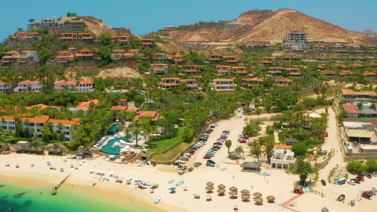Aerial shoreline with resort buildings, umbrellas, and hillside homes.