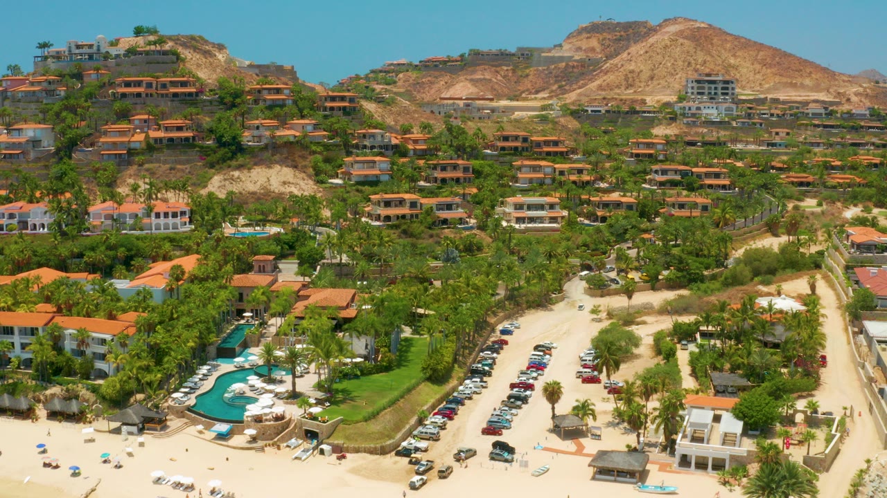 Aerial view of red roof buildings, pools, trees, roads, beach, and dry hills.