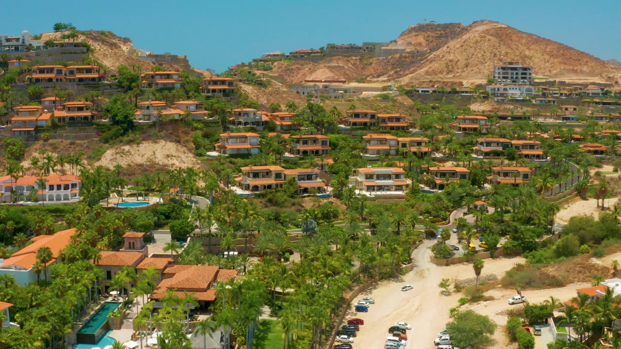 Opening frame of Palmilla Beach, Baja California Sur showing terracotta-roof buildings among palms, pale roads, a small blue pool, dry hills, blue sky, and no visible people.