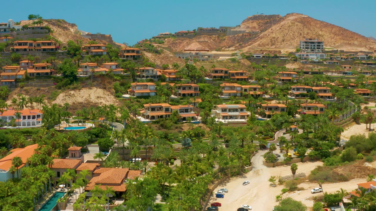 Third frame of Palmilla Beach, Baja California Sur showing clustered terracotta roofs, palms, a curving road, a small pool left of center, dry hills, and no visible people.