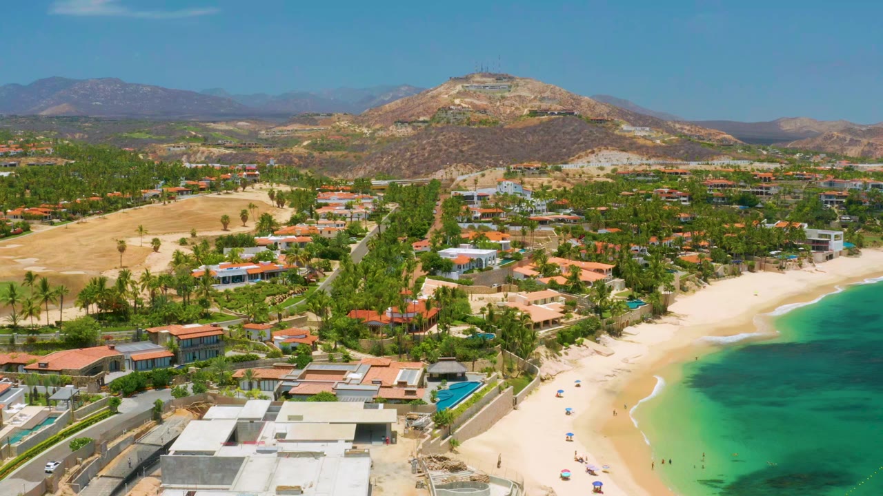 Opening aerial frame over Palmilla Beach in Baja California Sur, with sandy shoreline, clear green-blue water, beachfront homes, palms, dry hills, distant mountains, and no visible people.