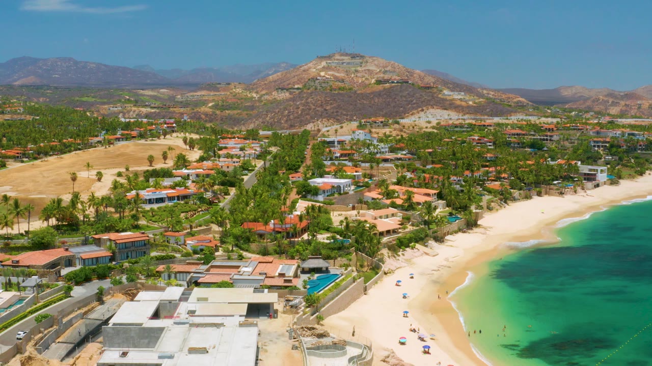 Palmilla Beach aerial frame with pale sand and green-blue water on the right, beachfront residences and palms through the center, arid hills beyond, and no visible people.
