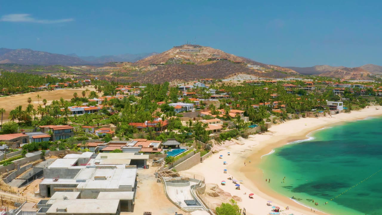 Aerial view of Palmilla Beach with turquoise water filling the right side, pale sand and parked cars along the shore, low buildings in front, and no visible people.