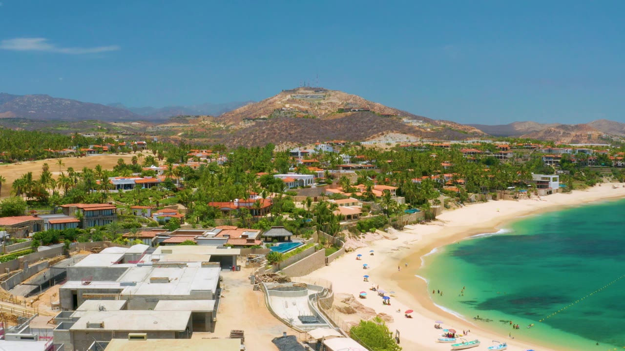 Elevated view of beachfront roofs, green palms, sandy beach, blue water, and hills.