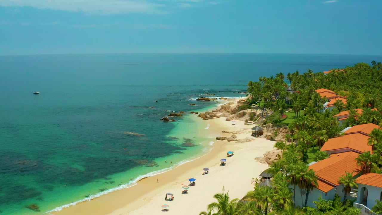 Opening aerial frame at Palmilla Beach in Baja California Sur showing blue water, pale sand, palm trees, rocky shoreline, orange-roofed houses, and no visible people.