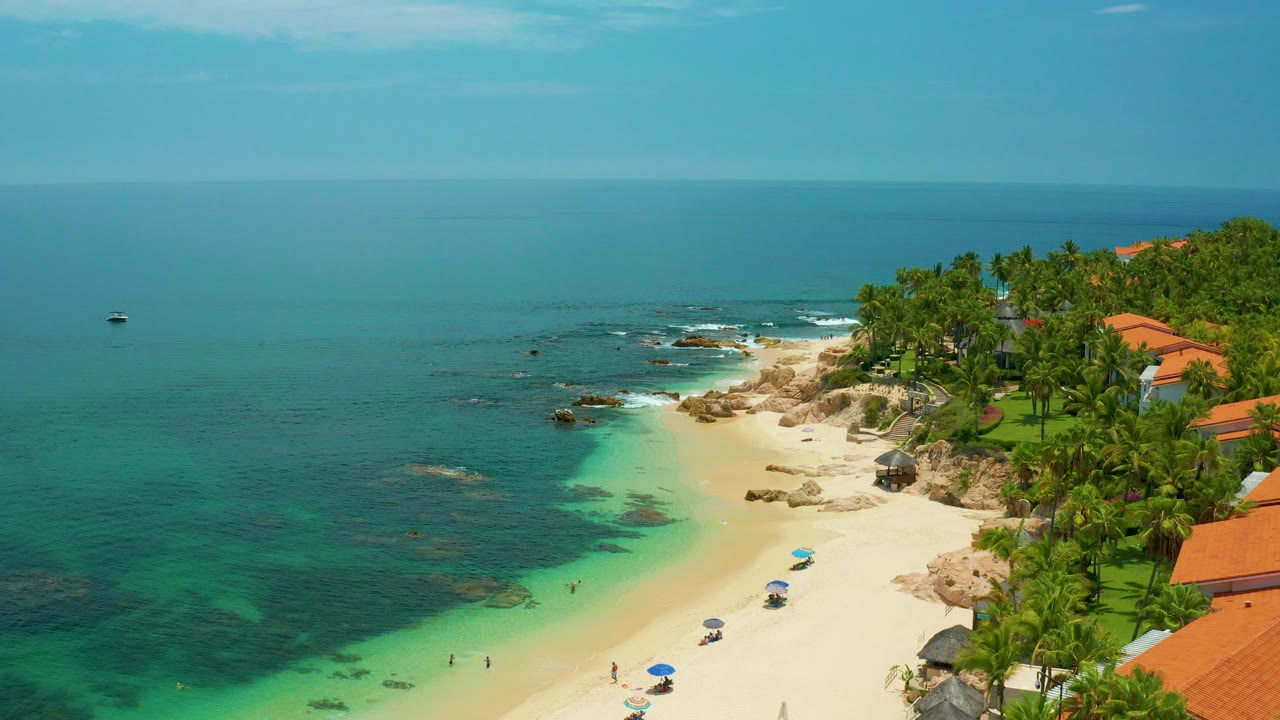 Aerial view over Palmilla Beach in Baja California Sur, Mexico, with turquoise water on the left, white sand below, palms and orange rooftops to the right, and no visible people.
