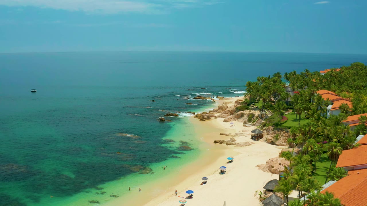 Wide aerial beach view with shallow green water, surf, palms, and rooftops.