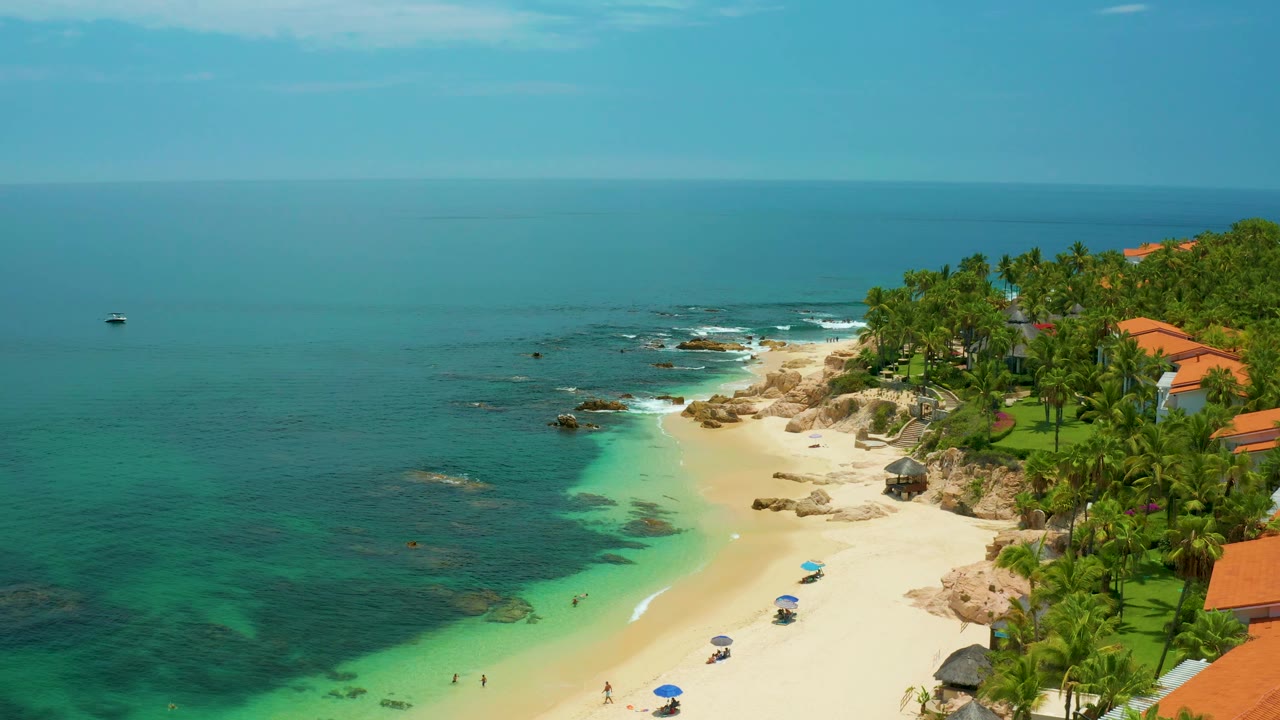 Aerial view at Palmilla Beach with rocky points near the surf, a white sand cove, palm-lined grounds, orange-roofed buildings, and no visible people.