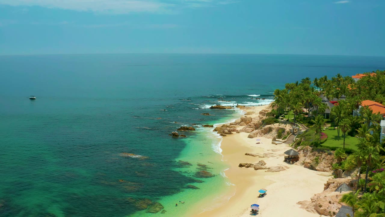 Aerial view one second into the Palmilla Beach clip, showing clear green-blue water around rocks, a curving sand beach, resort palms, and no visible people.