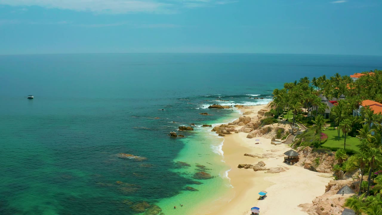 Palmilla Beach aerial frame with the open ocean across the left side, surf breaking around rocks, sand below, palms and red rooflines at the right, and no visible people.