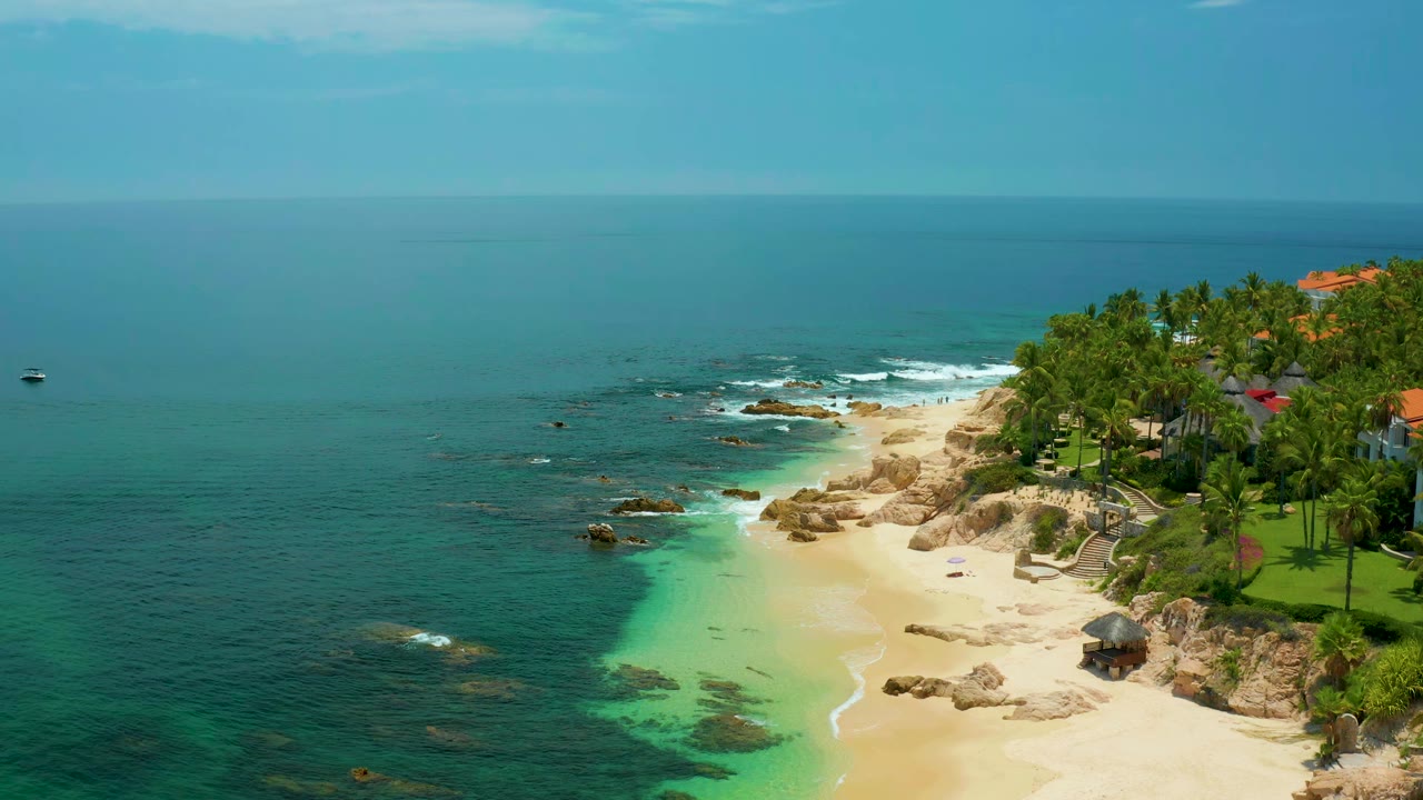 Wide aerial frame of Palmilla Beach showing deeper sea on the left, green-tinted shallow water near the sand, rock clusters along the shoreline, palms on the right edge, and no visible people.