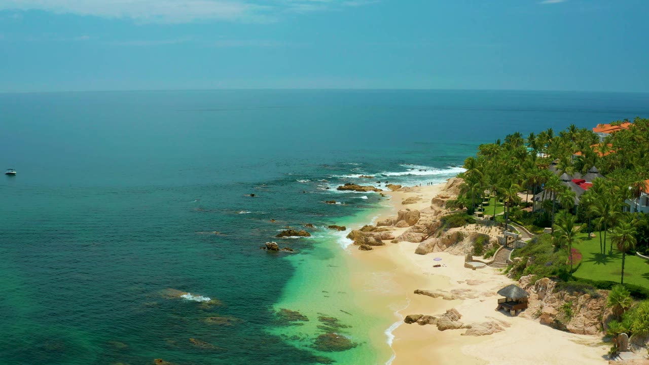 Aerial view at Palmilla Beach with the sandy cove pushed toward the right side, dark rocks in the shallows, small white waves offshore, dense palms, rooflines near the upper right, and no visible people.