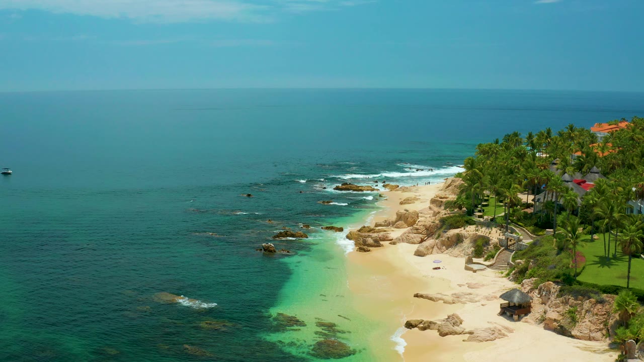 Aerial shoreline view with rocks, white surf, sand, palms, and blue water.