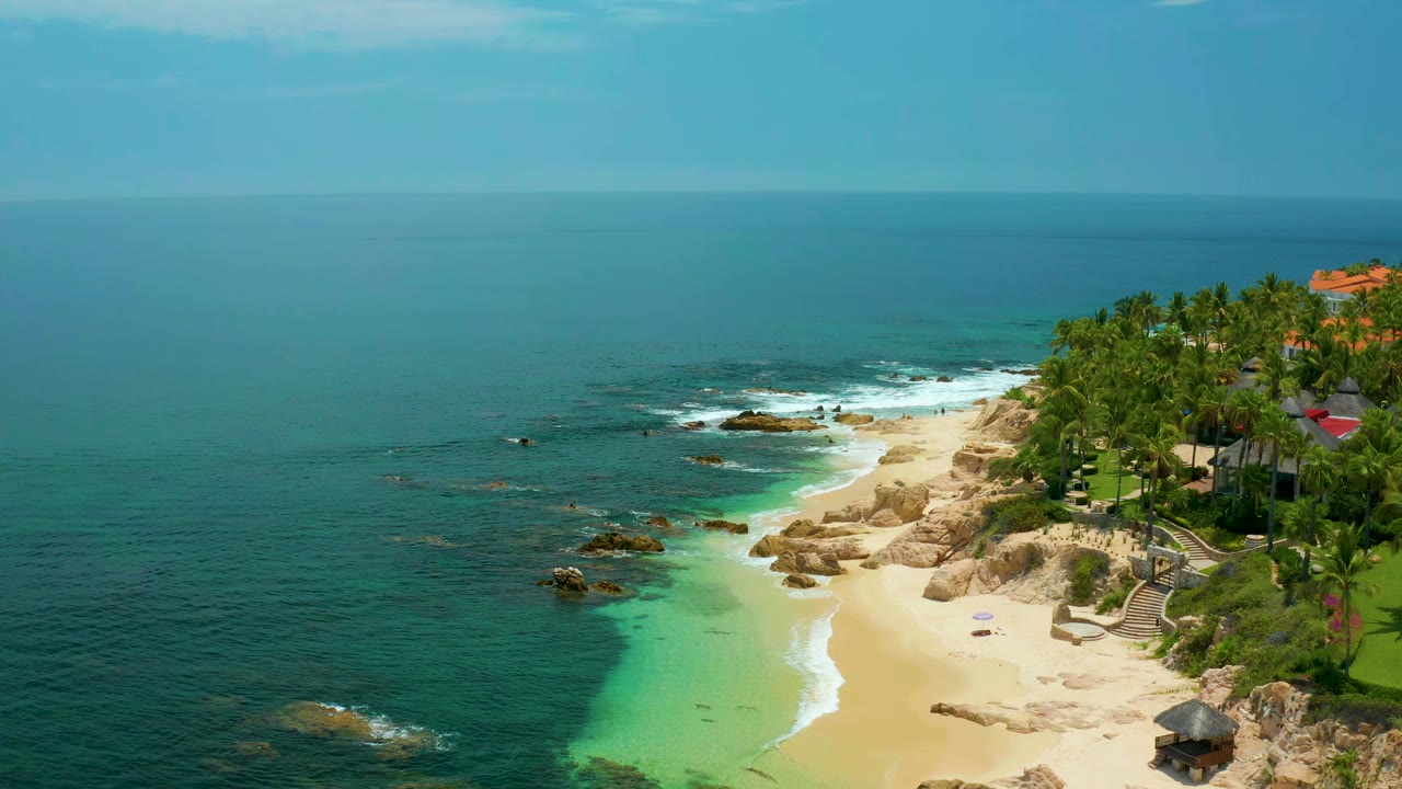 High coastal view of blue-green water meeting a sandy rocky beach and palms.
