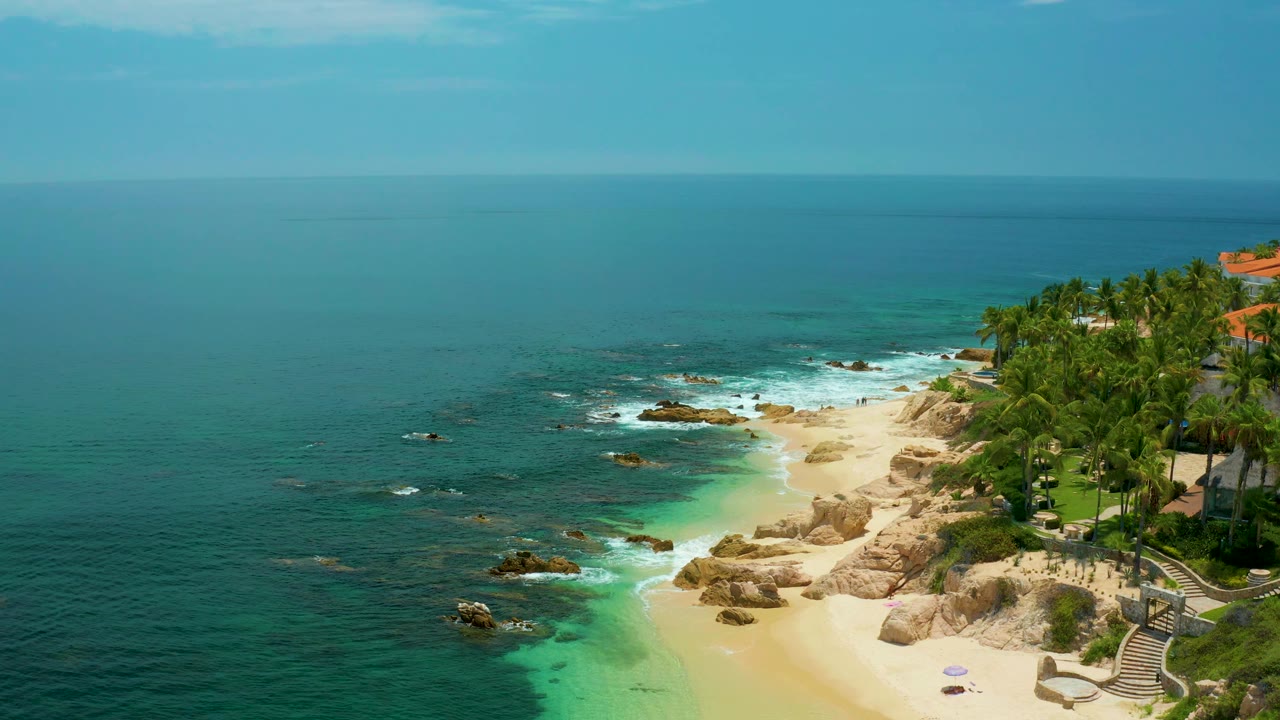Palmilla Beach aerial with teal shallows bright against darker ocean, pale sand and tan rocks through the center, dense palms along the right edge, red tile roofs above them, and no visible people.
