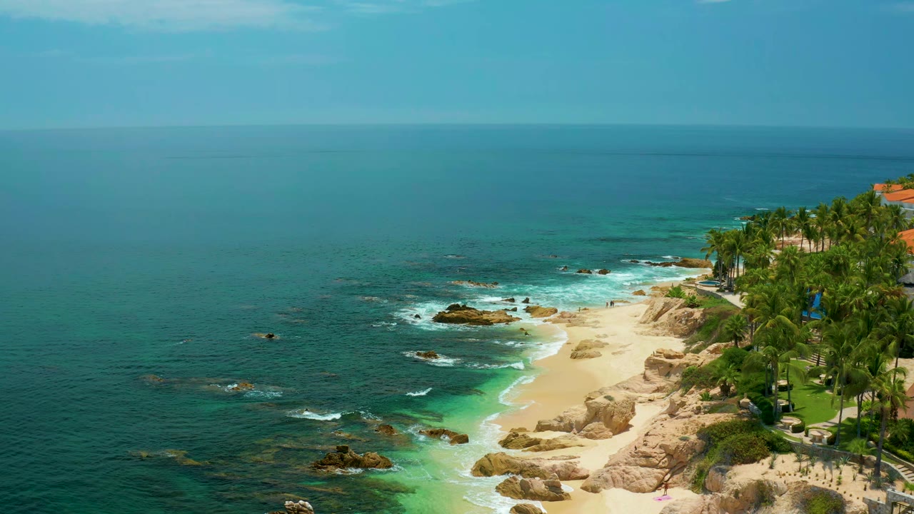 Wide aerial frame of Palmilla Beach with open blue water across the horizon, waves breaking around offshore rocks, sand below, palms on the right, and no visible people.