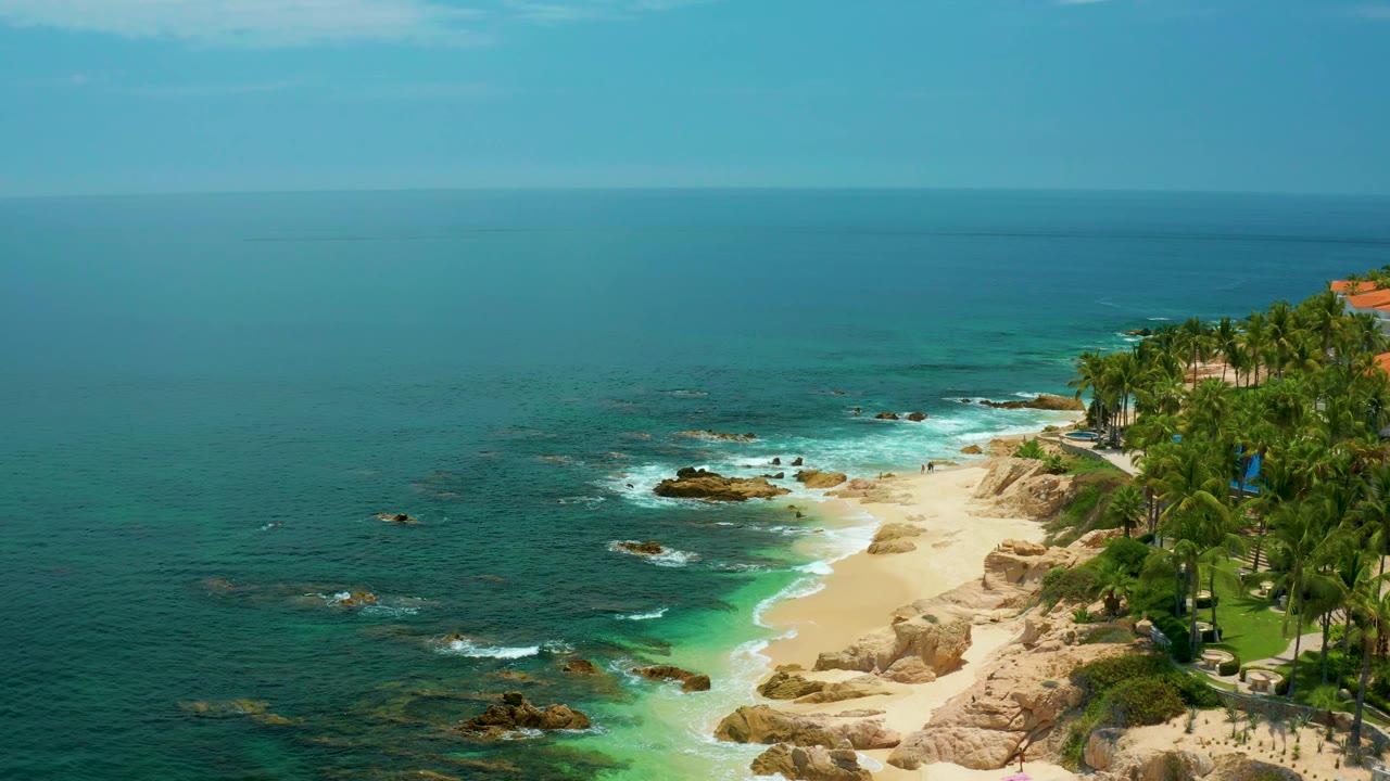 Aerial view of the Palmilla Beach shoreline with blue-green water, a bright strip of sand, surf curling along the beach, palms and greenery to the right, and no visible people.