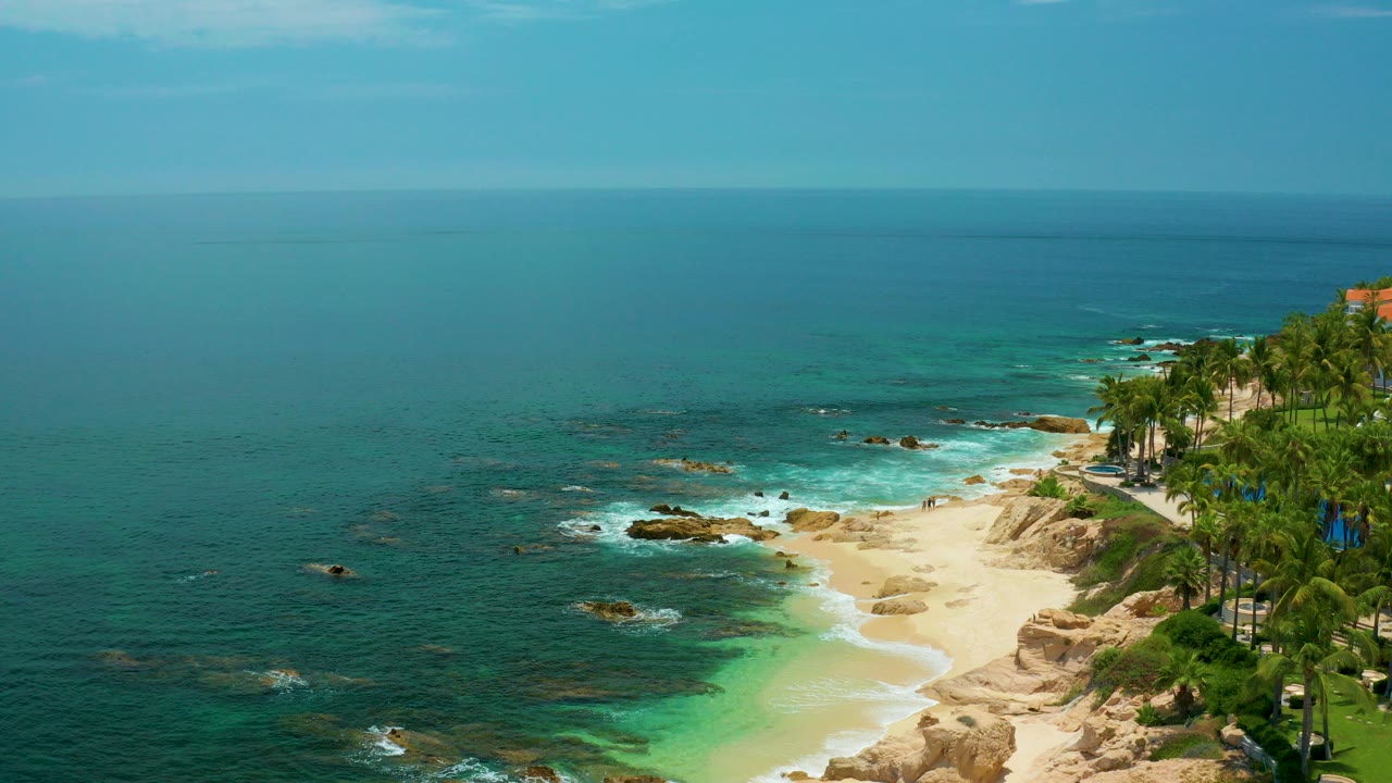 Wide aerial view of Palmilla Beach in Baja California Sur with turquoise water, pale sand, tan rocks, palms at the right edge, and no visible people.