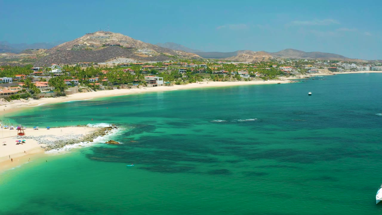 Opening aerial frame over Palmilla Beach, Baja California Sur, Mexico, with turquoise water in front, a sandy beach at left, shoreline homes below the hills, and no visible people.