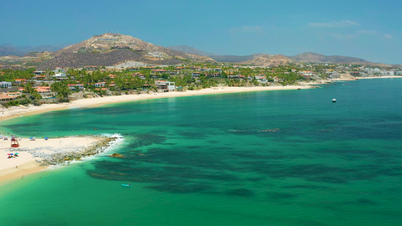 Palmilla Beach aerial view one second in, showing green-blue water bands, a pale beach curve, shoreline buildings, distant mountains, and no visible people.