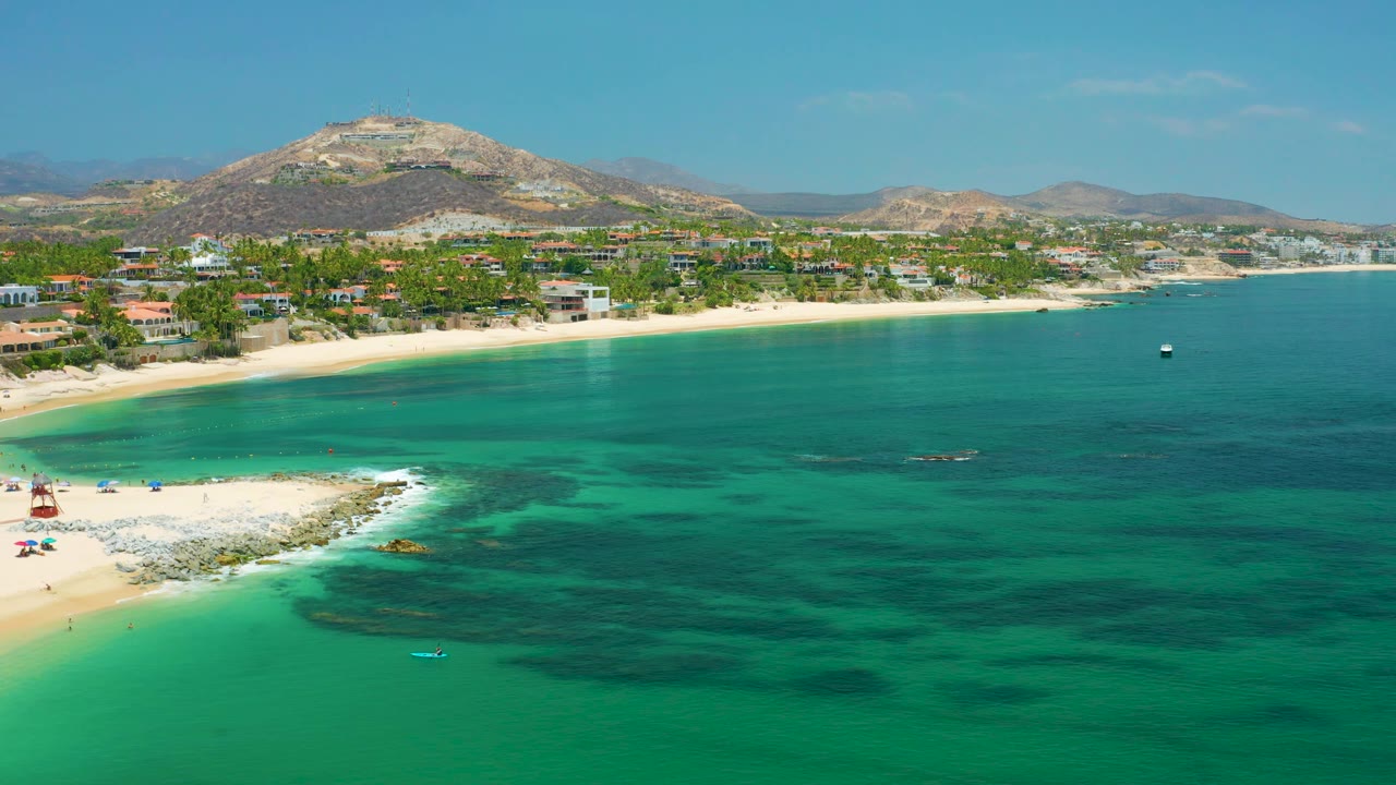 Palmilla Beach frame with the beach and rocky point low left, open turquoise water to the right, low coastal development, mountain ridges, and no visible people.