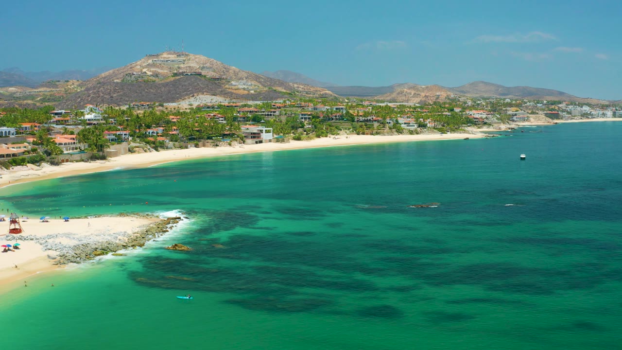 Beach aerial with white sand, clear water, shoreline buildings, and mountain backdrop.