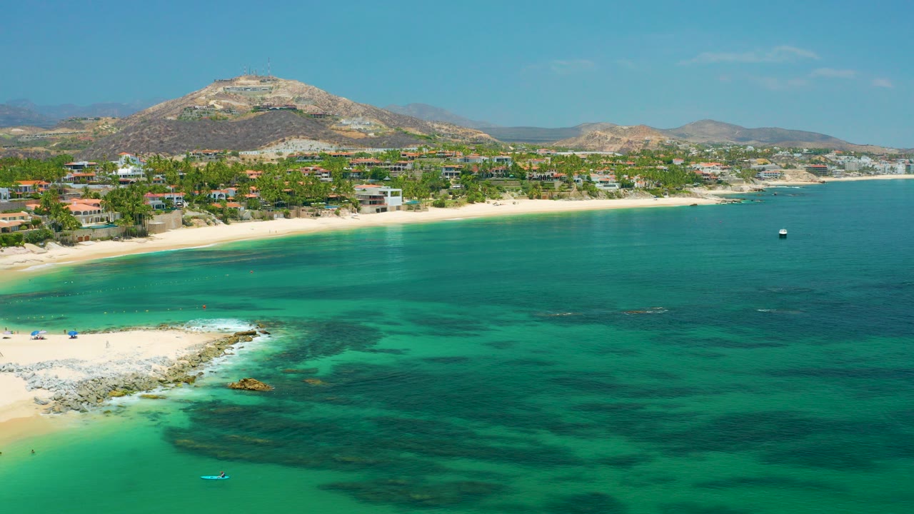 Opening aerial frame over Palmilla Beach, Baja California Sur, with turquoise ocean in front, a pale beach and shoreline homes beyond, dry hills on the horizon, and no visible people.