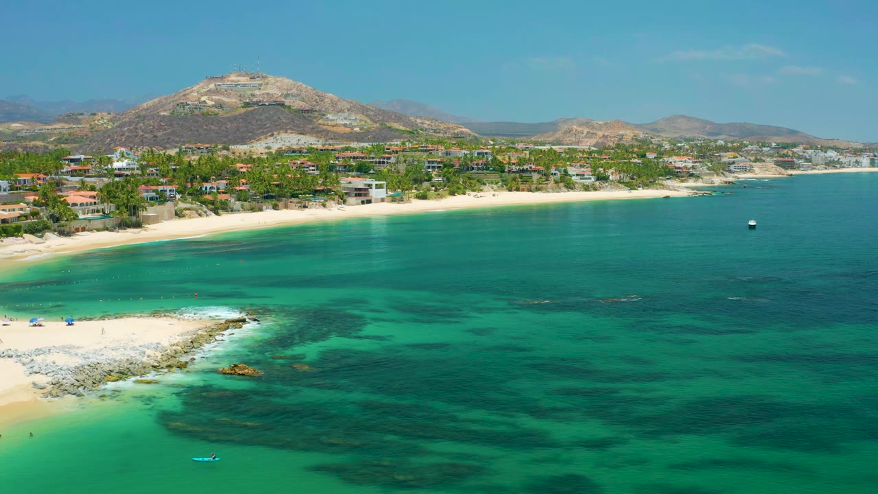 Wide drone view of clear coastal water, beach, homes, palms, and hills.