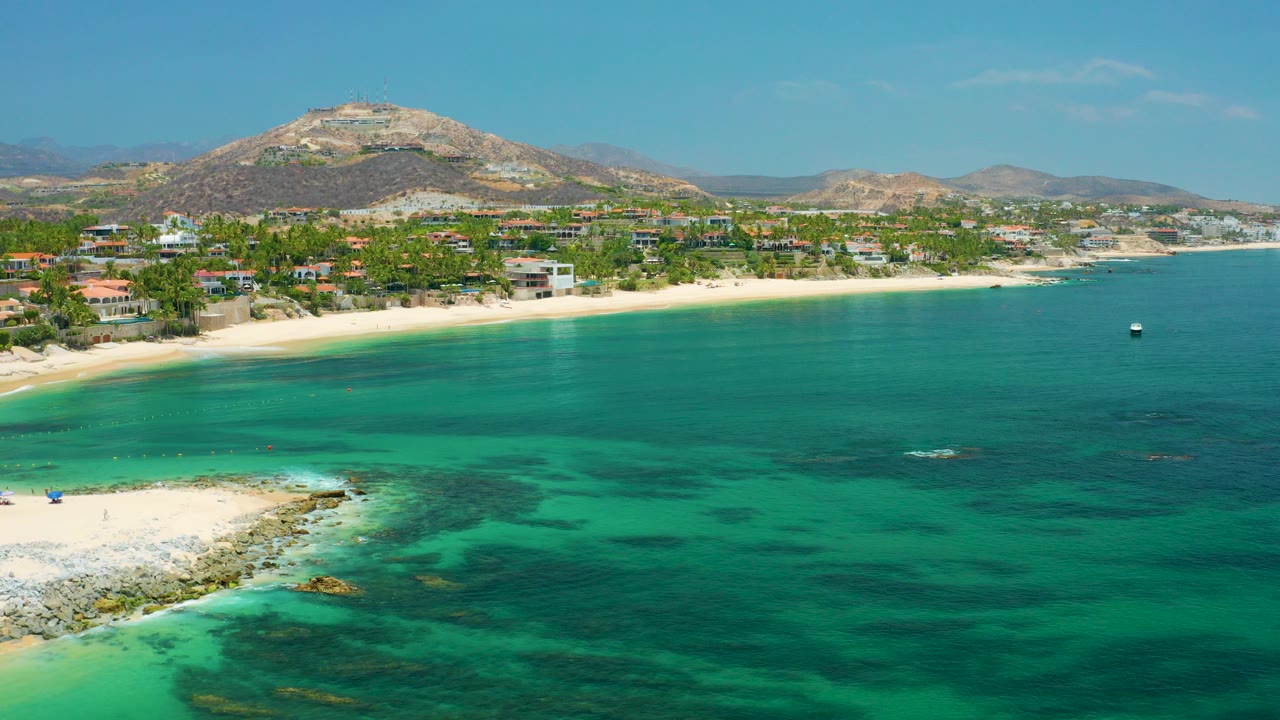 Palmilla Beach, Baja California Sur aerial view with turquoise water in front, a pale beach and hillside residences across the shore, dry mountains behind, and no visible people.
