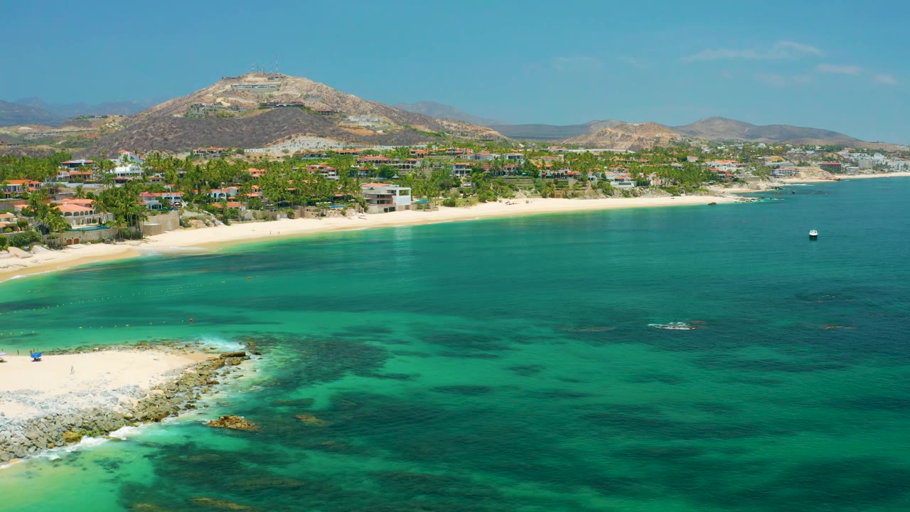 Palmilla Beach in Baja California Sur seen from above with clear teal water, dark reef patches, a long sand strip, hillside buildings, a small boat offshore, and no visible people.
