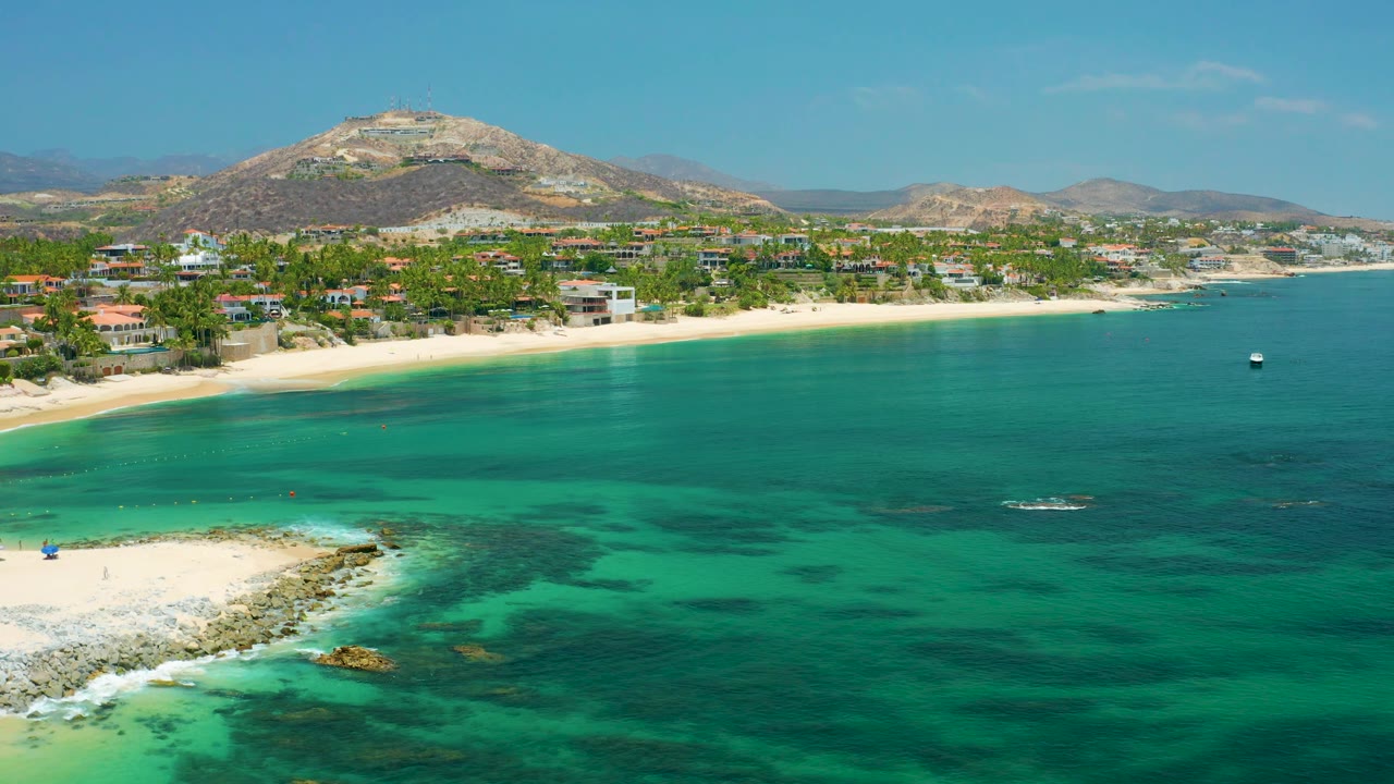 Aerial view over shallow water toward beach homes and a mountain ridge.