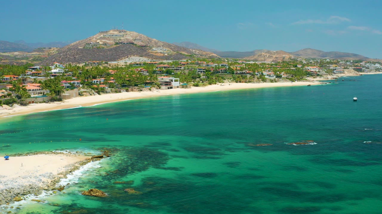 Opening aerial frame over Palmilla Beach, Baja California Sur, with turquoise water, dark reef patches, a sandy curve, shoreline buildings, dry hills, and no visible people.