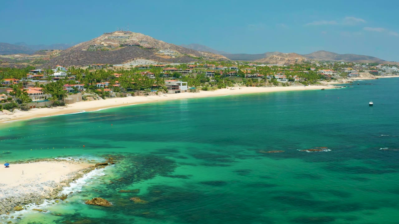 Wide aerial coastline with shallow blue-green water and beach homes below hills.