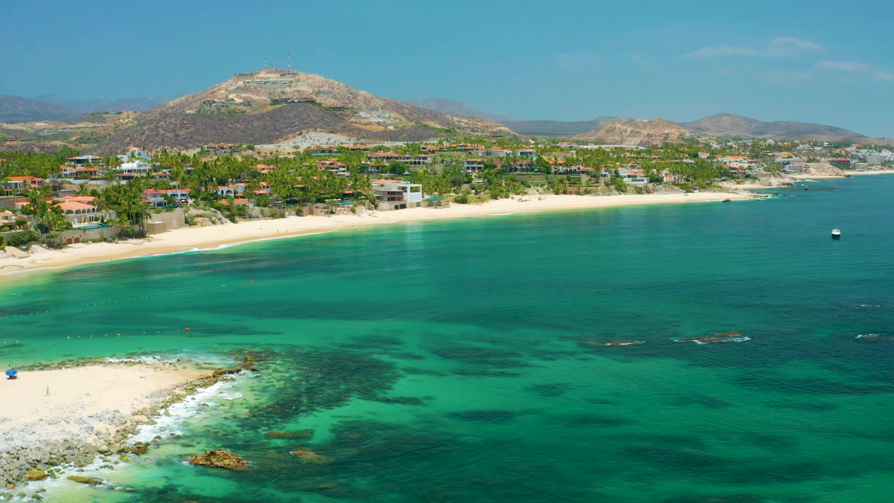 Palmilla Beach aerial view with reef-darkened turquoise water across the foreground, a rocky point at lower left, beach and low buildings along shore, and no visible people.