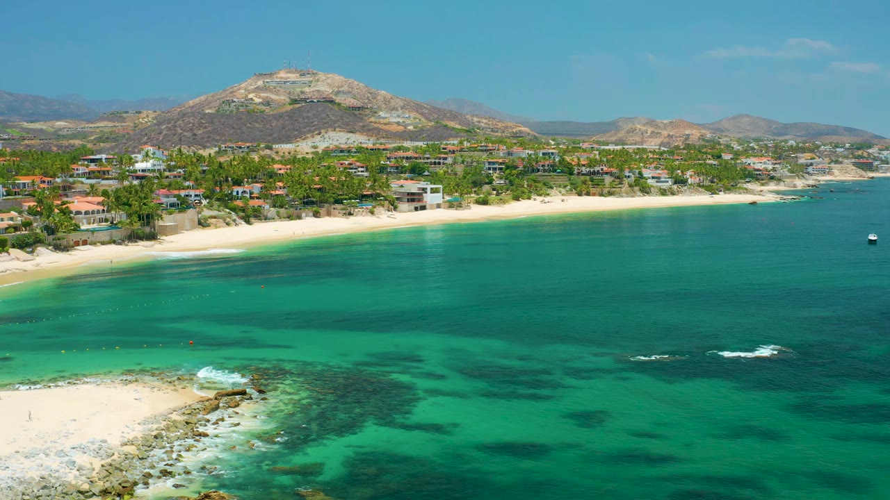 Palmilla Beach shoreline from above with a rocky foreground point, green-blue water, a palm-lined beach, scattered beachfront buildings, hills beyond, and no visible people.