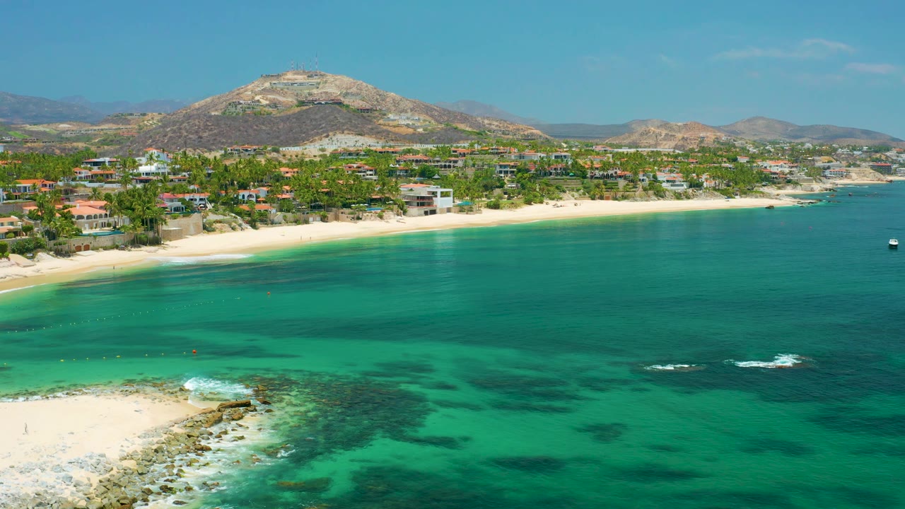 A wide view of Palmilla Beach, Baja California Sur, showing smooth turquoise water, a pale shoreline, palms and buildings along the coast, distant hills, and no visible people.